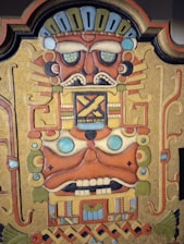 A detailed, colorful artwork depicting a stylized, ancient mask or face with geometric patterns. The face has large, expressive eyes with a vibrant design, a broad nose, and a mouth with distinct teeth. The piece is surrounded by intricate patterns and lines that add to the complexity and artistic style.
