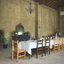 A rustic interior with a long wooden dining table covered with a white tablecloth and surrounded by mixed wooden chairs. The table is set with bottles, glasses, and plates, ready for a meal. In the background, an antique sideboard holds potted plants and a large mirror. The walls are rough, with a vintage chandelier hanging from the ceiling.