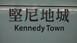 The image features a sign with Chinese characters above the text 'Kennedy Town'. The background is a muted teal color, and the text is in bold, black lettering.