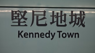 The image features a sign with Chinese characters above the text 'Kennedy Town'. The background is a muted teal color, and the text is in bold, black lettering.