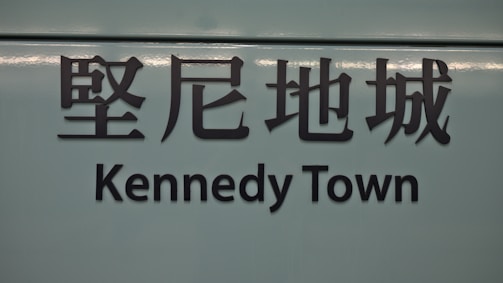 The image features a sign with Chinese characters above the text 'Kennedy Town'. The background is a muted teal color, and the text is in bold, black lettering.