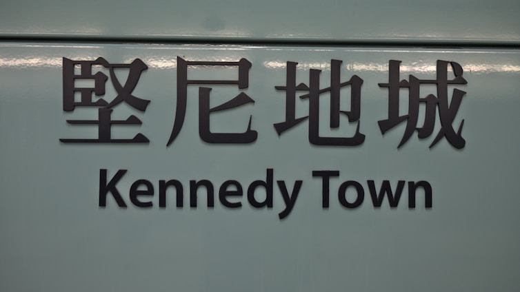 The image features a sign with Chinese characters above the text 'Kennedy Town'. The background is a muted teal color, and the text is in bold, black lettering.