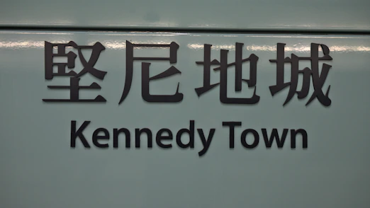 The image features a sign with Chinese characters above the text 'Kennedy Town'. The background is a muted teal color, and the text is in bold, black lettering.
