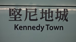 The image features a sign with Chinese characters above the text 'Kennedy Town'. The background is a muted teal color, and the text is in bold, black lettering.
