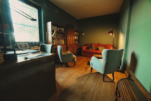 A cozy, warmly-lit living room featuring a wooden floor, a red sofa with cushions, two light blue armchairs, a wooden desk with a lamp and computer, and a bookshelf filled with books. A large window allows natural light to spill into the room, complementing the ambient lighting provided by a floor lamp.