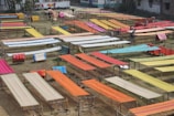 Various brightly colored fabrics are laid out on elevated wooden frames in an open outdoor space, possibly for drying or display. The area is organized with multiple rows of textiles, showcasing colors like orange, red, yellow, and blue. Some individuals are seen working among the fabrics.