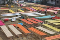 Various brightly colored fabrics are laid out on elevated wooden frames in an open outdoor space, possibly for drying or display. The area is organized with multiple rows of textiles, showcasing colors like orange, red, yellow, and blue. Some individuals are seen working among the fabrics.