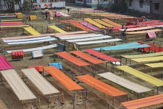 Various brightly colored fabrics are laid out on elevated wooden frames in an open outdoor space, possibly for drying or display. The area is organized with multiple rows of textiles, showcasing colors like orange, red, yellow, and blue. Some individuals are seen working among the fabrics.