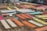 Various brightly colored fabrics are laid out on elevated wooden frames in an open outdoor space, possibly for drying or display. The area is organized with multiple rows of textiles, showcasing colors like orange, red, yellow, and blue. Some individuals are seen working among the fabrics.