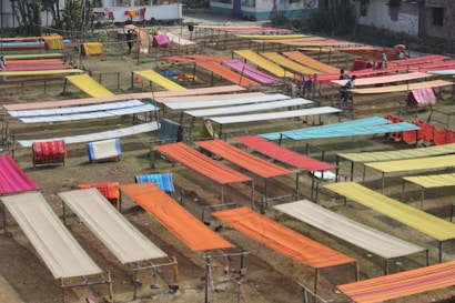 Various brightly colored fabrics are laid out on elevated wooden frames in an open outdoor space, possibly for drying or display. The area is organized with multiple rows of textiles, showcasing colors like orange, red, yellow, and blue. Some individuals are seen working among the fabrics.