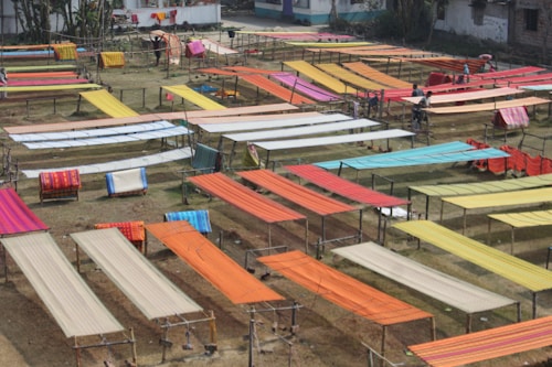 Various brightly colored fabrics are laid out on elevated wooden frames in an open outdoor space, possibly for drying or display. The area is organized with multiple rows of textiles, showcasing colors like orange, red, yellow, and blue. Some individuals are seen working among the fabrics.