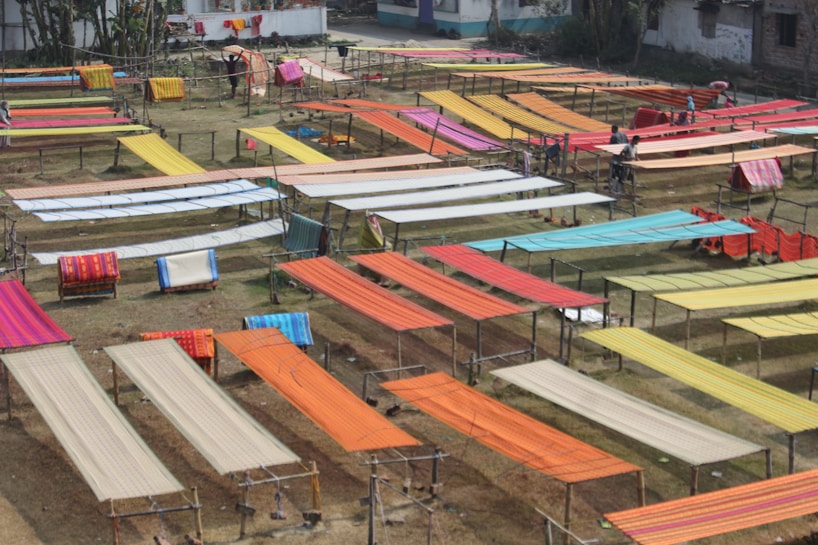 Various brightly colored fabrics are laid out on elevated wooden frames in an open outdoor space, possibly for drying or display. The area is organized with multiple rows of textiles, showcasing colors like orange, red, yellow, and blue. Some individuals are seen working among the fabrics.