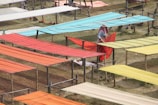 Colorful fabric pieces are laid out on wooden frames in an outdoor setting for drying or display. A person wearing a hat and headscarf is adjusting a piece of red fabric. The rows of fabrics in shades of orange, red, yellow, and blue create a vibrant pattern against a grassy background.