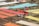 Colorful fabric pieces are laid out on wooden frames in an outdoor setting for drying or display. A person wearing a hat and headscarf is adjusting a piece of red fabric. The rows of fabrics in shades of orange, red, yellow, and blue create a vibrant pattern against a grassy background.