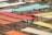 Colorful fabric pieces are laid out on wooden frames in an outdoor setting for drying or display. A person wearing a hat and headscarf is adjusting a piece of red fabric. The rows of fabrics in shades of orange, red, yellow, and blue create a vibrant pattern against a grassy background.