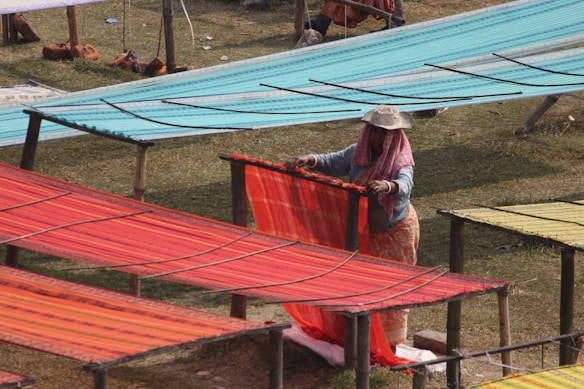 A person is arranging colorful fabric panels on wooden racks in an outdoor setting. Each panel displays vibrant patterns, with predominant colors including shades of red, orange, and teal. The person is dressed in modest clothing and wears a hat to shield from the sun, suggesting a warm climate.