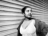A woman stands confidently with her hand on her hip, leaning against a corrugated metal shutter. She wears a lace top and a coat draped over her shoulders. Her expression is thoughtful as she gazes to the side.