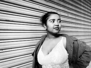 A woman stands confidently with her hand on her hip, leaning against a corrugated metal shutter. She wears a lace top and a coat draped over her shoulders. Her expression is thoughtful as she gazes to the side.