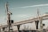 A large industrial area featuring several cranes near a port. A modern bridge with a complex steel structure stretches across the scene in the background. The sky has a muted tone, adding to the industrial aesthetic.