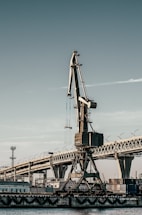 A large industrial crane stands prominently against a backdrop of a bridge and several buildings. The crane's metal structure is detailed, and it appears to be positioned at a dockside or port. The bridge features curving barriers with multiple struts.