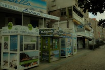 A street view featuring several ticket booths advertising trips to the Isla de Ons. The booths are adorned with various travel posters. Nearby, there are buildings with signs indicating restaurants, including one named 'Titanic Marisquería'. The atmosphere appears quiet, and the cobblestone pavement adds a rustic charm to the scene.