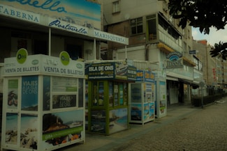 A street view featuring several ticket booths advertising trips to the Isla de Ons. The booths are adorned with various travel posters. Nearby, there are buildings with signs indicating restaurants, including one named 'Titanic Marisquer&iacute;a'. The atmosphere appears quiet, and the cobblestone pavement adds a rustic charm to the scene.