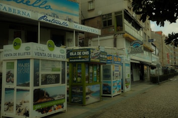 A street view featuring several ticket booths advertising trips to the Isla de Ons. The booths are adorned with various travel posters. Nearby, there are buildings with signs indicating restaurants, including one named 'Titanic Marisquer&iacute;a'. The atmosphere appears quiet, and the cobblestone pavement adds a rustic charm to the scene.
