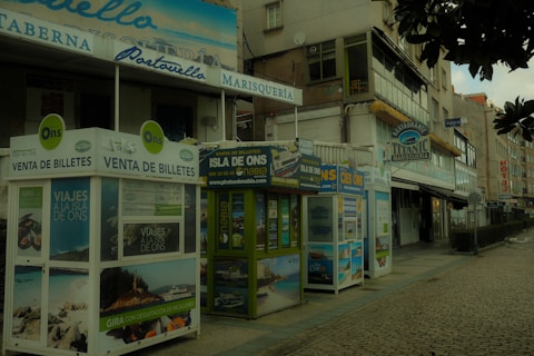 A street view featuring several ticket booths advertising trips to the Isla de Ons. The booths are adorned with various travel posters. Nearby, there are buildings with signs indicating restaurants, including one named 'Titanic Marisquer&iacute;a'. The atmosphere appears quiet, and the cobblestone pavement adds a rustic charm to the scene.