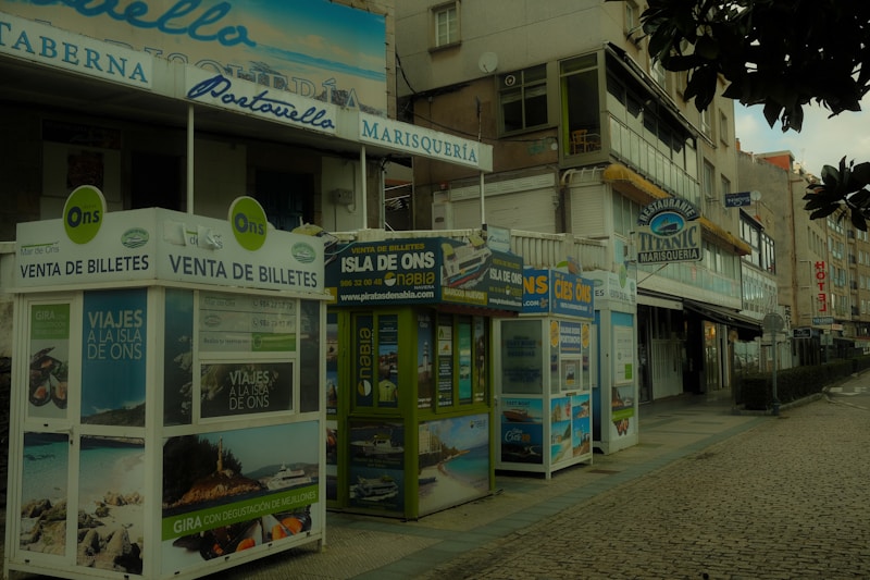 A street view featuring several ticket booths advertising trips to the Isla de Ons. The booths are adorned with various travel posters. Nearby, there are buildings with signs indicating restaurants, including one named 'Titanic Marisquería'. The atmosphere appears quiet, and the cobblestone pavement adds a rustic charm to the scene.