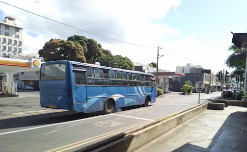 A blue bus is driving on an empty street in a city area. In the background, there is a Shell gas station with a tree partially obscuring its signage. Buildings with multiple stories are visible on the left, under a slightly cloudy sky.