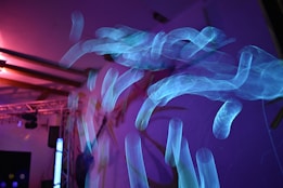 Abstract light patterns create an ethereal and dynamic effect with swirling blue shapes against a dark background, accentuated by pink and purple lighting. The setting appears to be a dimly lit room or stage, suggesting movement and artistic expression.
