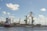 A large cargo ship is docked at an industrial port. The ship is marked with the name 'WAGENBORG' and appears to be loaded with green containers. Several large yellow cranes are positioned over the ship for loading and unloading purposes. The port facilities include warehouses and other industrial buildings, and there is a partly cloudy sky above.