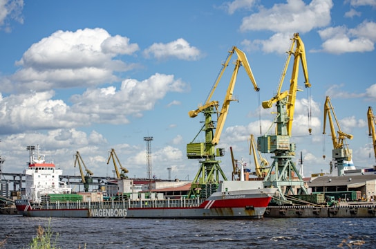 A cargo ship is docked at a busy industrial port. Several large yellow cranes are positioned along the pier, towering over the ship and various buildings. The sky is partly cloudy with a few patches of blue visible.