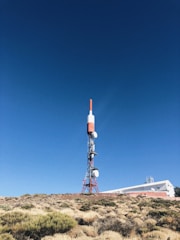 A tall communication tower stands in a clear, blue sky. The structure is primarily red and white, with several satellite dishes attached. It is situated on a rocky, bush-covered hillside next to a white building with a flat roof, partially visible in the background.