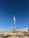 A tall communication tower stands in a clear, blue sky. The structure is primarily red and white, with several satellite dishes attached. It is situated on a rocky, bush-covered hillside next to a white building with a flat roof, partially visible in the background.