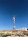 A tall communication tower stands in a clear, blue sky. The structure is primarily red and white, with several satellite dishes attached. It is situated on a rocky, bush-covered hillside next to a white building with a flat roof, partially visible in the background.