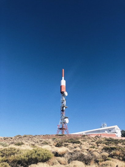 A tall communication tower stands in a clear, blue sky. The structure is primarily red and white, with several satellite dishes attached. It is situated on a rocky, bush-covered hillside next to a white building with a flat roof, partially visible in the background.
