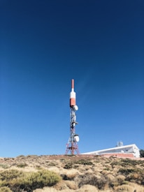 A tall communication tower stands in a clear, blue sky. The structure is primarily red and white, with several satellite dishes attached. It is situated on a rocky, bush-covered hillside next to a white building with a flat roof, partially visible in the background.