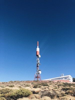 A tall communication tower stands in a clear, blue sky. The structure is primarily red and white, with several satellite dishes attached. It is situated on a rocky, bush-covered hillside next to a white building with a flat roof, partially visible in the background.
