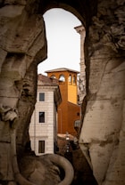 An architectural scene viewed through a stone archway, revealing a mix of historic and rustic buildings. The central building is painted in a vibrant orange, contrasting with the surrounding neutral-toned structures. Stone sculptures and carvings frame the view, adding an artistic touch.