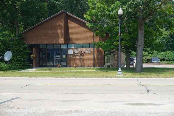 A small building serves as the United States Post Office for Caseville, MI, with a modern architectural design featuring a wooden exterior and large glass windows. It is situated next to a road with trees and greenery surrounding the area. A satellite dish is visible on the left side, and there are two parked cars partially visible on the right.