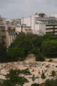 Aerial view of an urban park featuring a circular playground with colorful play equipment like slides and climbers. Surrounding the playground are trees and large buildings, creating a contrast between green space and urban architecture. Several people and children are visible, enjoying the recreational area.