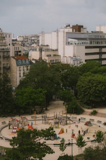 Aerial view of an urban park featuring a circular playground with colorful play equipment like slides and climbers. Surrounding the playground are trees and large buildings, creating a contrast between green space and urban architecture. Several people and children are visible, enjoying the recreational area.