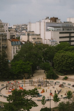 Aerial view of an urban park featuring a circular playground with colorful play equipment like slides and climbers. Surrounding the playground are trees and large buildings, creating a contrast between green space and urban architecture. Several people and children are visible, enjoying the recreational area.