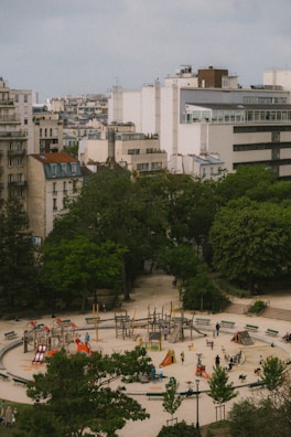 Aerial view of an urban park featuring a circular playground with colorful play equipment like slides and climbers. Surrounding the playground are trees and large buildings, creating a contrast between green space and urban architecture. Several people and children are visible, enjoying the recreational area.