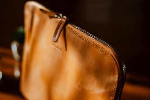 A close-up view of a brown leather pouch with a zipper. The leather appears smooth and well-crafted, highlighted by warm lighting that accentuates its texture. The zipper is slightly open, and the stitching around the edges is visible.