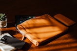A leather pouch and phone case are positioned on a wooden surface with a tablet partially visible underneath. A smartphone and a spiral notebook with a black pen are nearby. Sunlight casts soft shadows across the items, adding warmth to the scene. A small succulent plant in a glass pot is also present, contributing a touch of greenery.
