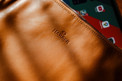 A close-up of a brown leather pouch with a visible zipper, partially covering a tablet displaying various app icons. The leather has a smooth texture with the brand name embossed. The lighting casts soft shadows across the surface.