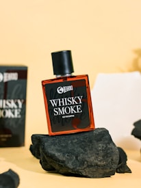 A bottle of perfume labeled 'Whisky Smoke' by Beardo is prominently displayed on a dark, textured rock. The background has a soft yellow hue and another box of the same perfume is partially visible. The scene conveys a rugged and sophisticated atmosphere.