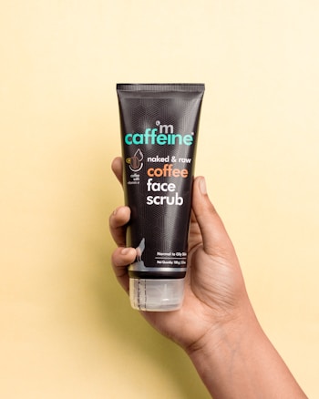 A hand is holding a tube of mCaffeine naked & raw coffee face scrub against a light yellow background. The tube is black with geometric patterns and features the product name in bold letters.